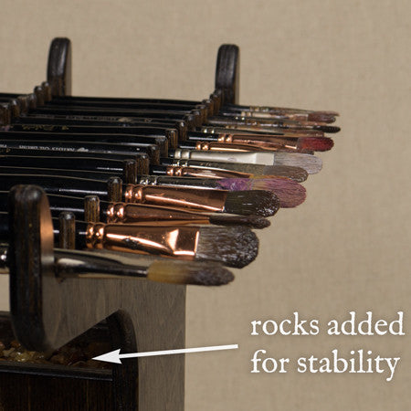 Paintbrushes resting in the Geneva Brush Holder, with a note that rocks are added for stability